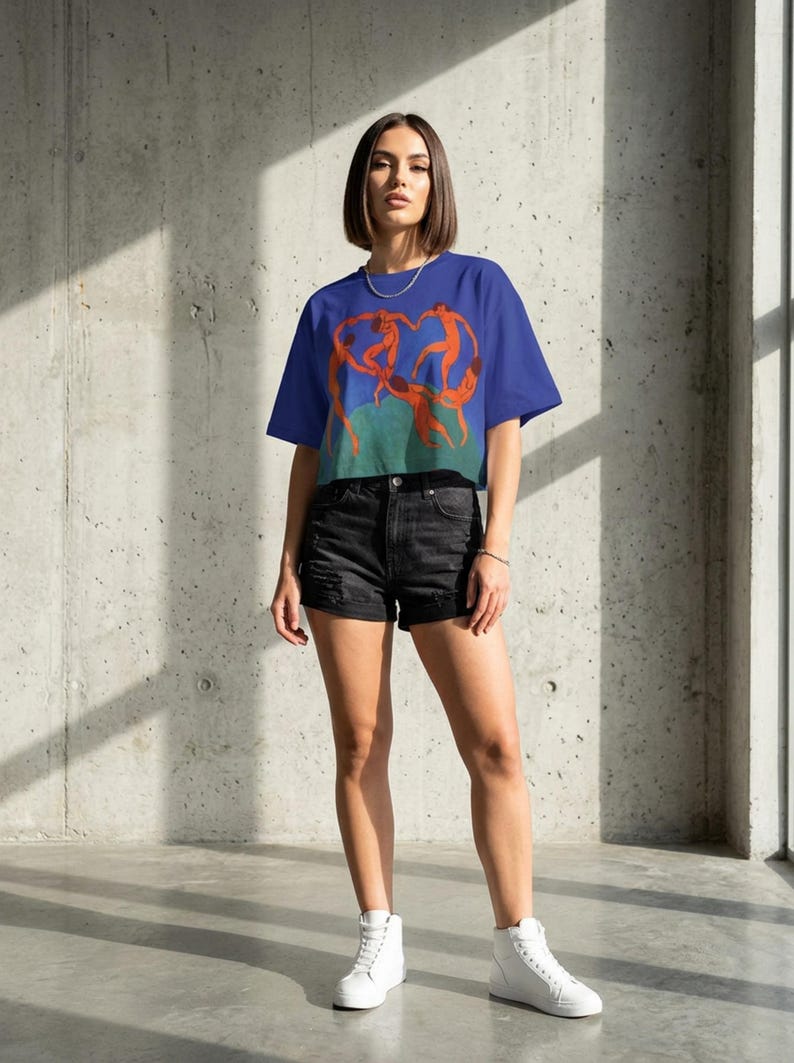 May include: A blue crop top with an orange illustration of dancing figures. The model is wearing black denim shorts and white high-top trainers. The background is a concrete wall.