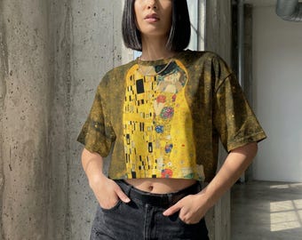 Puede incluir: Una camiseta corta con un estampado de la obra "El Beso" de Gustav Klimt. La camiseta tiene una combinación de colores amarillo y dorado, con un aspecto desgastado. La modelo lleva unos shorts vaqueros negros.