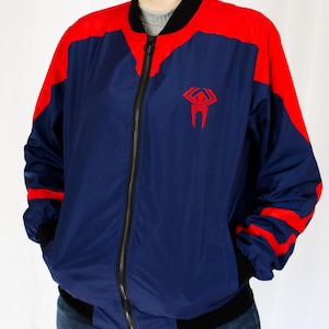 May include: A blue and red bomber jacket with a red spider logo on the front. The jacket has a zipper closure and black ribbed cuffs and waistband.