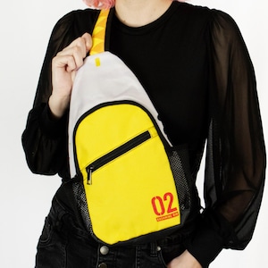 May include: A yellow and white sling bag with a black zipper and mesh pocket. The bag has the number "02" printed in red, with text below. The strap is yellow with a white accent. The bag is worn across the body.