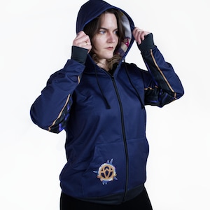 K&ouml;nnte beinhalten: Ein marineblauer Rei&szlig;verschluss-Hoodie mit einem goldenen und violetten Design an den &Auml;rmeln und einem goldenen Symbol auf der Vorderseite. Der Hoodie hat eine Kapuze mit einem dunkelvioletten Futter.