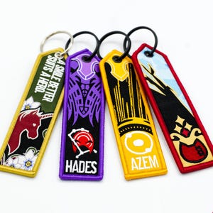 May include: Four embroidered keyrings with black rings. The keyrings feature various designs and text, including a unicorn, the word "HADES", and the word "AZEM". The keyrings have a variety of colours, including yellow, purple, red, and green.