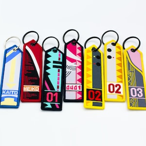 May include: Embroidered keychains in various colors, including blue, red, black, pink, and yellow. Each keychain features unique designs, text, and numbers. The keychains have black metal rings for attachment.