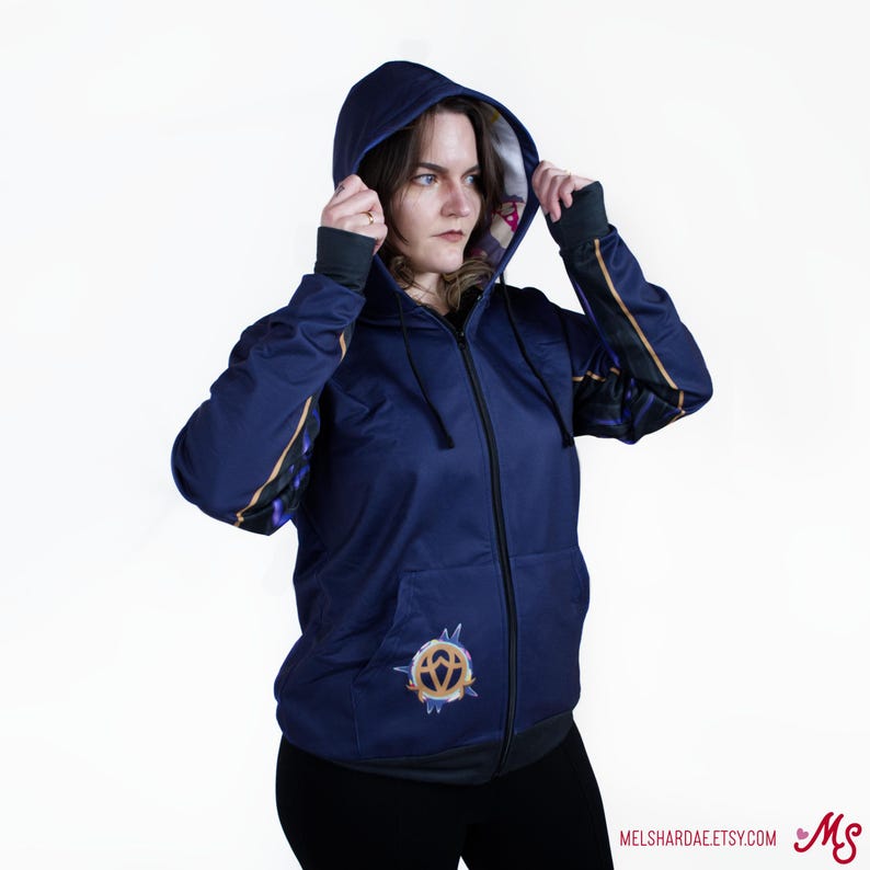 K&ouml;nnte beinhalten: Ein marineblauer Rei&szlig;verschluss-Hoodie mit einem goldenen und violetten Design an den &Auml;rmeln und einem goldenen Symbol auf der Vorderseite. Der Hoodie hat eine Kapuze mit einem dunkelvioletten Futter.