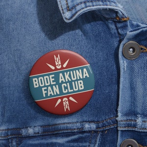 May include: A round button with the text "BODE AKUNA FAN CLUB" in white letters on a teal stripe, set against a red background. The button features white graphic designs and is pinned to a denim jacket.
