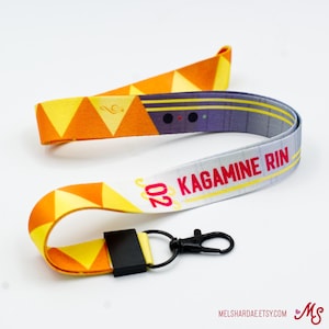 May include: A white, yellow, and orange lanyard with a black and gray pattern and the text "202 Kagamine Rin" printed on it. The lanyard has a black breakaway clasp and a silver metal hook.