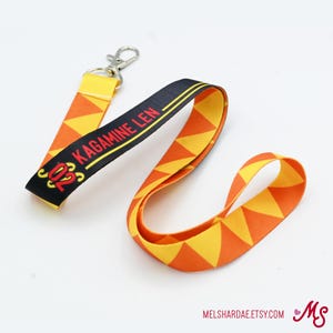 May include: A black and yellow lanyard with the text "2023 Kagamine Len" printed on it. The lanyard has a triangle pattern and a silver clip.