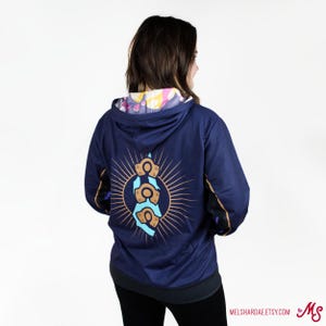 K&ouml;nnte beinhalten: Ein marineblauer Hoodie mit einem bunten Design auf der Innenseite der Kapuze. Die R&uuml;ckseite des Hoodies zeigt eine Grafik eines blauen und goldenen Symbols mit einem Sonnenstrahlendesign.
