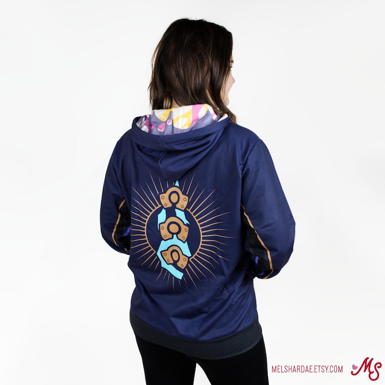 K&ouml;nnte beinhalten: Ein marineblauer Hoodie mit einem bunten Design auf der Innenseite der Kapuze. Die R&uuml;ckseite des Hoodies zeigt eine Grafik eines blauen und goldenen Symbols mit einem Sonnenstrahlendesign.