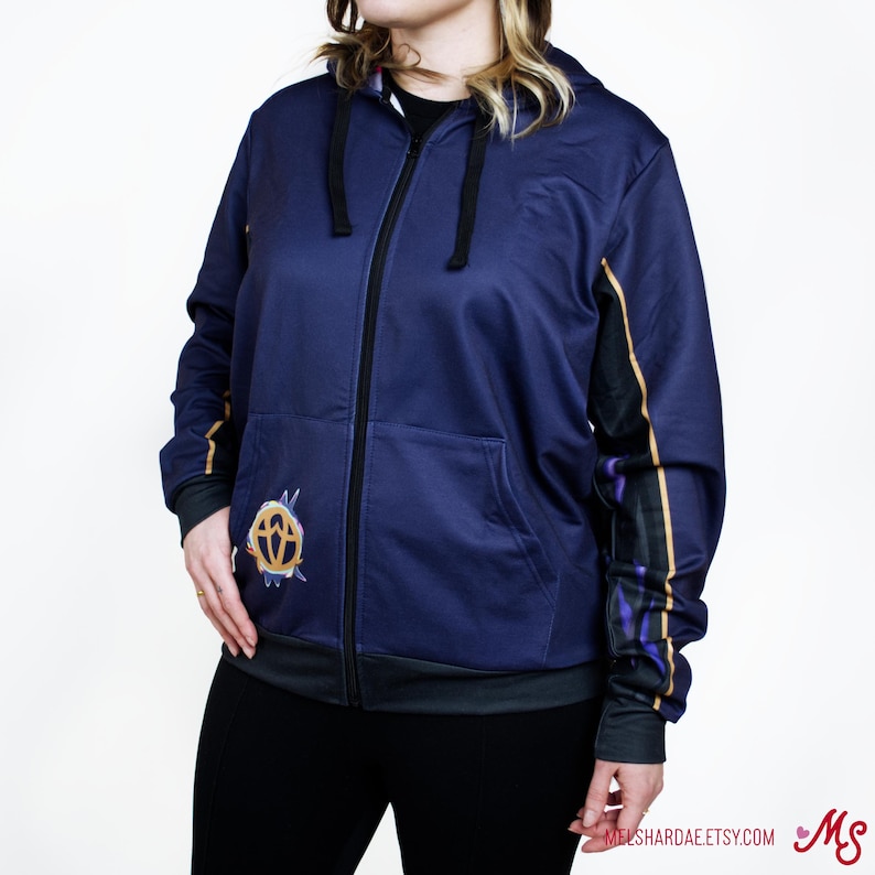 K&ouml;nnte beinhalten: Ein marineblauer Rei&szlig;verschluss-Hoodie mit einem goldenen und violetten Design an den &Auml;rmeln und einem goldenen Symbol auf der Vorderseite. Der Hoodie hat einen schwarzen Rei&szlig;verschluss und einen schwarzen Kordelzug.