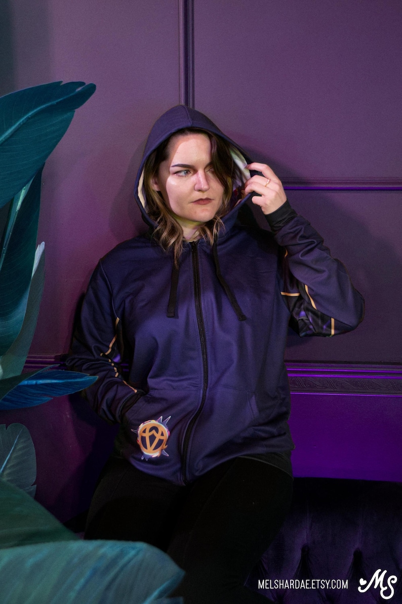 K&ouml;nnte beinhalten: Ein dunkelblauer Kapuzenpullover mit einem goldenen Design auf der Vorderseite. Der Hoodie hat eine Kapuze und einen Rei&szlig;verschluss. Das Design auf der Vorderseite ist ein stilisiertes Symbol mit einer Diamantenform und einem Kreis darum.