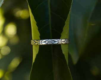 May include: A silver ring with a delicate floral design is nestled between two green leaves.
