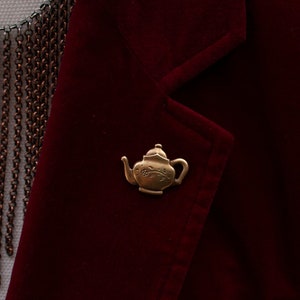 Può includere: Una spilla a forma di teiera dorata con un motivo floreale su un blazer color borgogna.