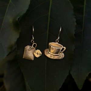 May include: Gold-tone earrings with a teacup and tea bag charm. The earrings are hanging from a green leafy branch.