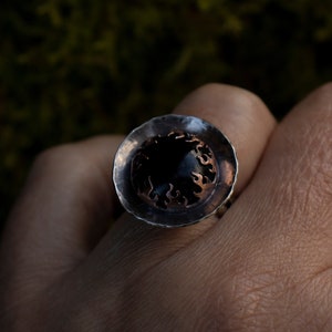 Puede incluir: Un anillo artesanal con una piedra grande, oscura y redonda engastada en metal plateado. El anillo tiene un diseño único con un patrón en forma de llama de color cobre alrededor de la piedra. Es una joya.