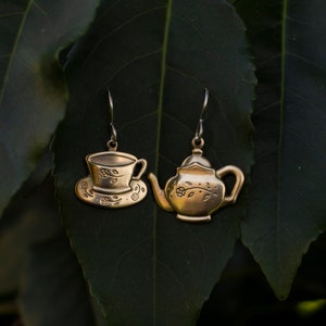 Puede incluir: Un par de pendientes de color dorado con forma de tetera y taza de té con platillo. Los pendientes cuelgan de un fondo verde frondoso.