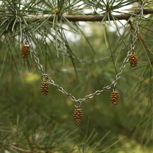 Puede incluir: Un collar de cadena plateada con cinco colgantes de piña de pino dorados. El collar está fotografiado sobre un fondo de ramas de pino verde.