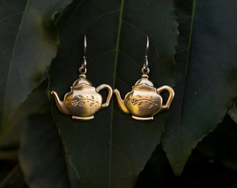 May include: A pair of gold teapot-shaped earrings with floral details hanging from a leafy green background.