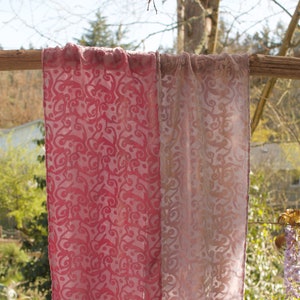 May include: Two sheer fabric panels with a pink and white ombre effect and a swirling pattern. The panels are hanging from a wooden branch and have fringe at the bottom.