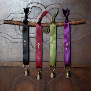 Peut inclure: Quatre marque-pages en ruban coloré, noir, bordeaux, vert olive et violet, ornés de breloques dorées en forme de clé et de petites cloches en laiton. Ils sont suspendus à une branche en bois.