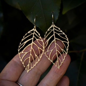 May include: A pair of gold leaf-shaped earrings with a delicate, intricate design. The earrings are made of metal and have a slightly textured finish.