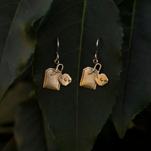 May include: A pair of gold-toned earrings shaped like tea bags with a small tag that says "tea" hanging from each. The earrings are hanging from a green leafy branch.