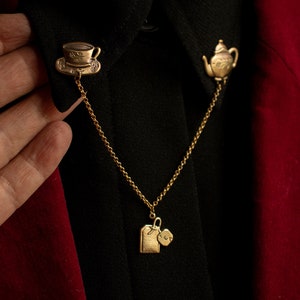 Puede incluir: Un collar de cadena dorada con tres colgantes: una taza de té, una tetera y una bolsita de té. Los colgantes están unidos a una camisa negra.