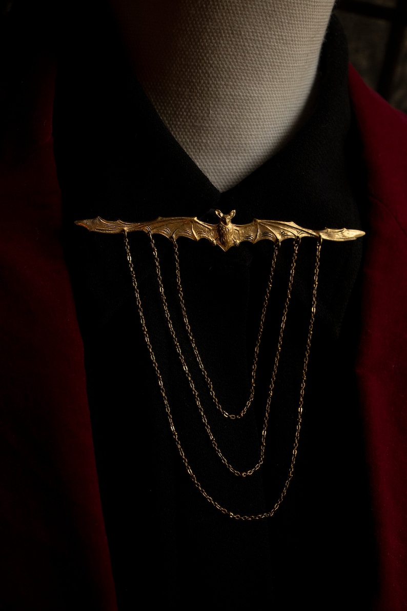 Pu&ograve; includere: Un fermaglio a forma di pipistrello dorato con un dettaglio a catena. Il fermaglio &egrave; indossato su una camicia nera con una giacca rossa.