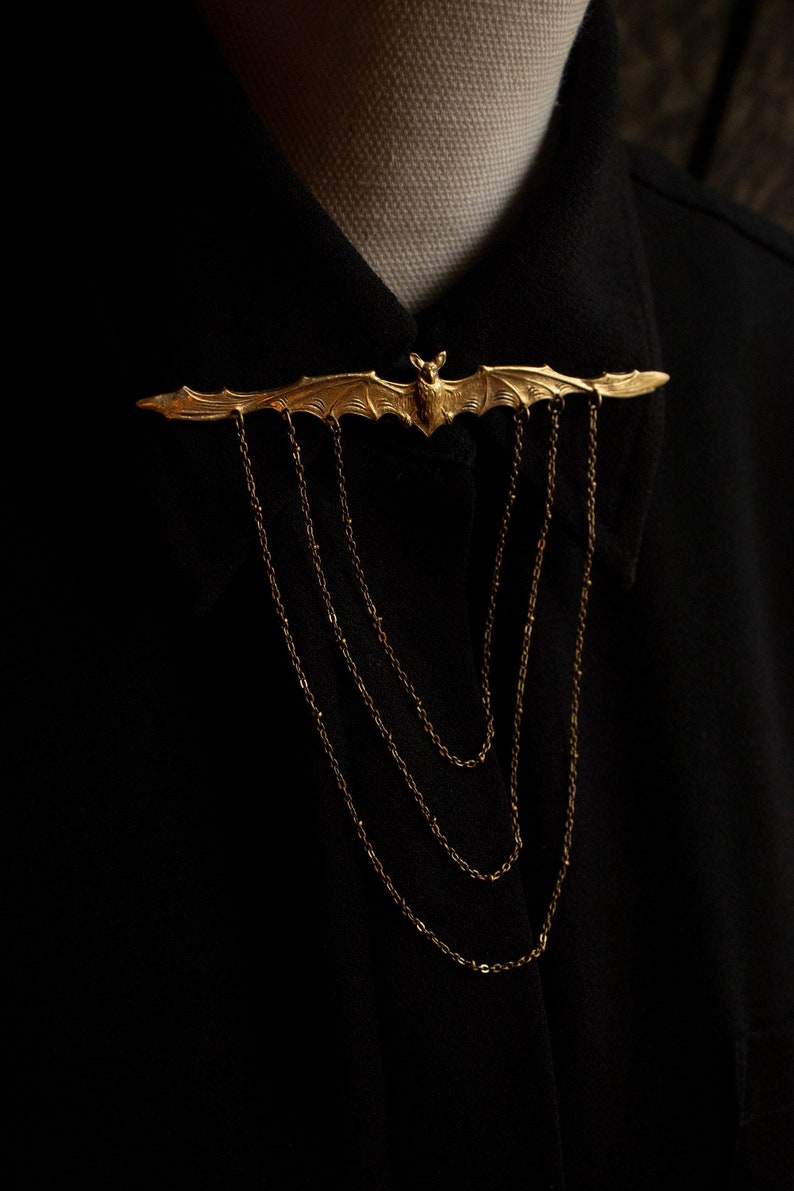 Pu&ograve; includere: Un fermaglio a forma di pipistrello dorato con catene pendenti. Il fermaglio &egrave; indossato su una camicia nera con colletto.
