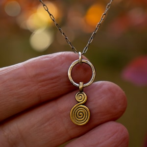 Puede incluir: Un collar de cadena de plata con un colgante de oro con un dise&ntilde;o de doble espiral. El colgante est&aacute; unido a un anillo de plata.