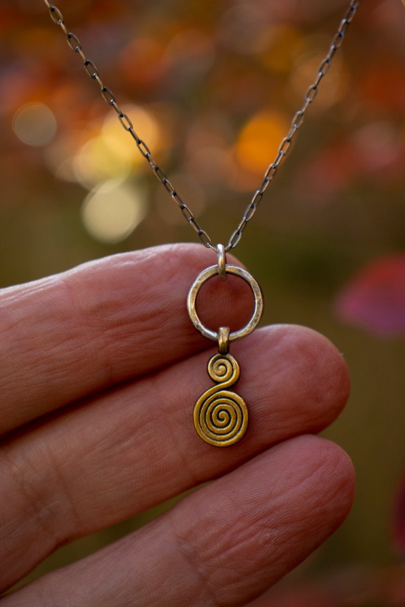 Puede incluir: Un collar de cadena de plata con un colgante de oro con un dise&ntilde;o de doble espiral. El colgante est&aacute; unido a un anillo de plata.