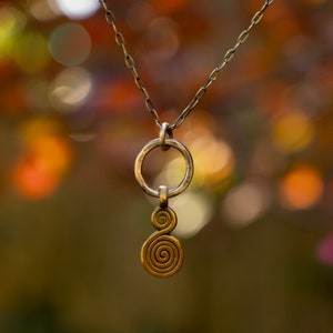 Puede incluir: Un collar de cadena dorada con un colgante que presenta un c&iacute;rculo y un dise&ntilde;o de doble espiral.