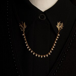May include: A gold chain necklace with white beads and lily flower accents. The necklace is worn on a black shirt with a button.