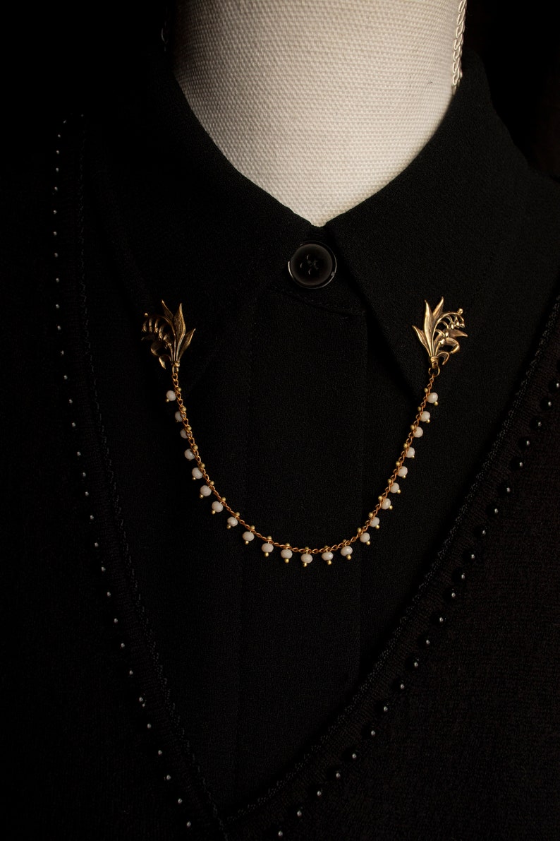 May include: A gold chain necklace with white beads and lily flower accents. The necklace is worn on a black shirt with a button.
