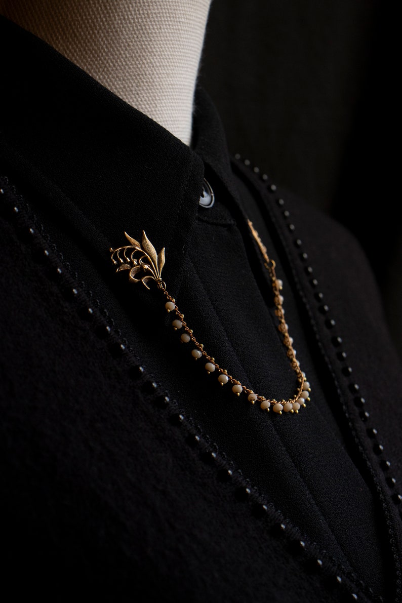 May include: A gold chain necklace with a lily flower charm and small white beads. The necklace is on a black shirt.