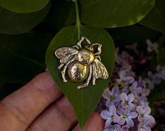 May include: A gold-colored bee pin with detailed wings and body, resting on a green leaf. The leaf is held by a hand, with purple lilac flowers in the background.