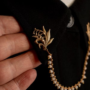 May include: A gold lily of the valley brooch with a chain of white beads attached. The brooch is pinned to a black fabric collar.