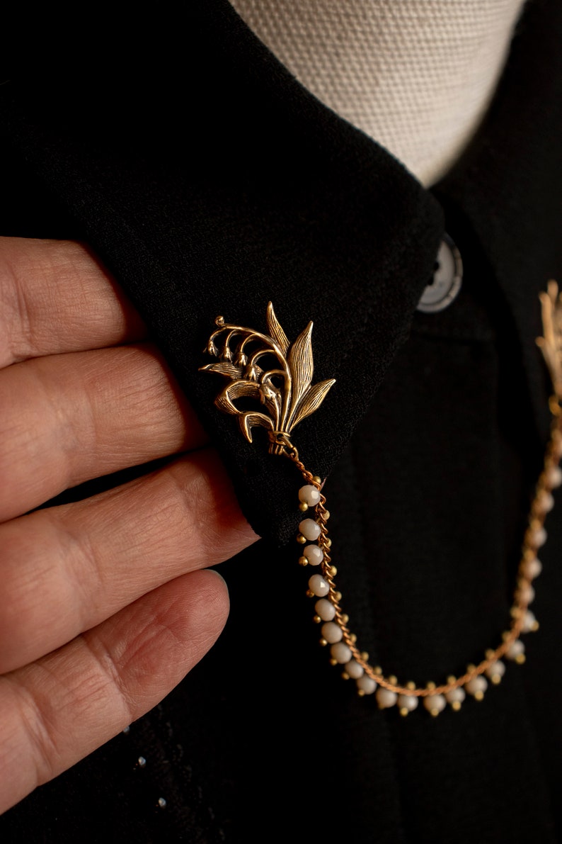 May include: A gold lily of the valley brooch with a chain of white beads attached. The brooch is pinned to a black fabric collar.
