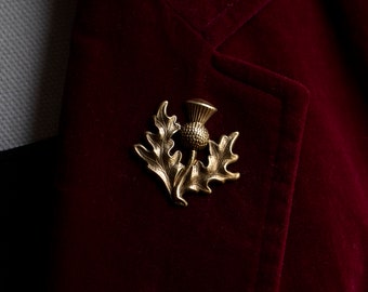 Puede incluir: Broche de cardo dorado prendido en una blazer de terciopelo burdeos. El broche presenta una flor de cardo detallada con hojas. La blazer tiene un diseño clásico.
