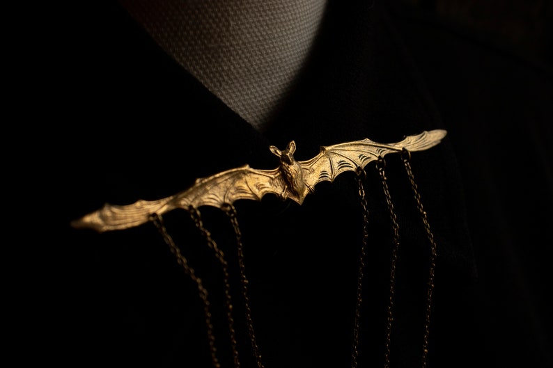 Pu&ograve; includere: Un fermaglio a forma di pipistrello dorato con catene pendenti. Il pipistrello &egrave; dettagliato con intricati motivi di ali e un corpo testurizzato.