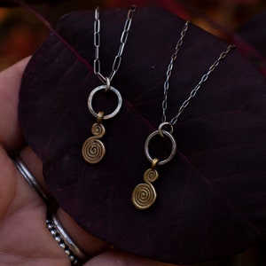 Puede incluir: Dos collares de cadena plateada con colgantes de oro. Los colgantes son un c&iacute;rculo con un dise&ntilde;o en espiral. Los collares est&aacute;n sobre una hoja de color rojo oscuro.