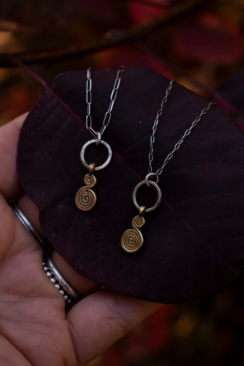 Puede incluir: Dos collares de cadena plateada con colgantes de oro. Los colgantes son un c&iacute;rculo con un dise&ntilde;o en espiral. Los collares est&aacute;n sobre una hoja de color rojo oscuro.