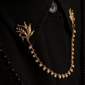May include: A gold chain necklace with a lily flower charm and white beads. The necklace is on a black fabric background.