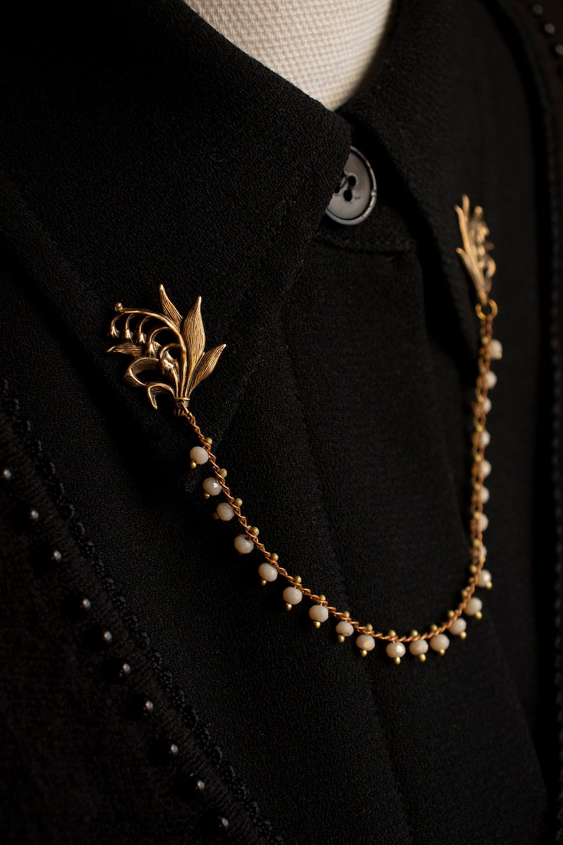 May include: A gold chain necklace with a lily flower charm and white beads. The necklace is on a black fabric background.
