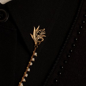 May include: A gold lily of the valley brooch with a chain of small white beads. The brooch is pinned to a black fabric background.