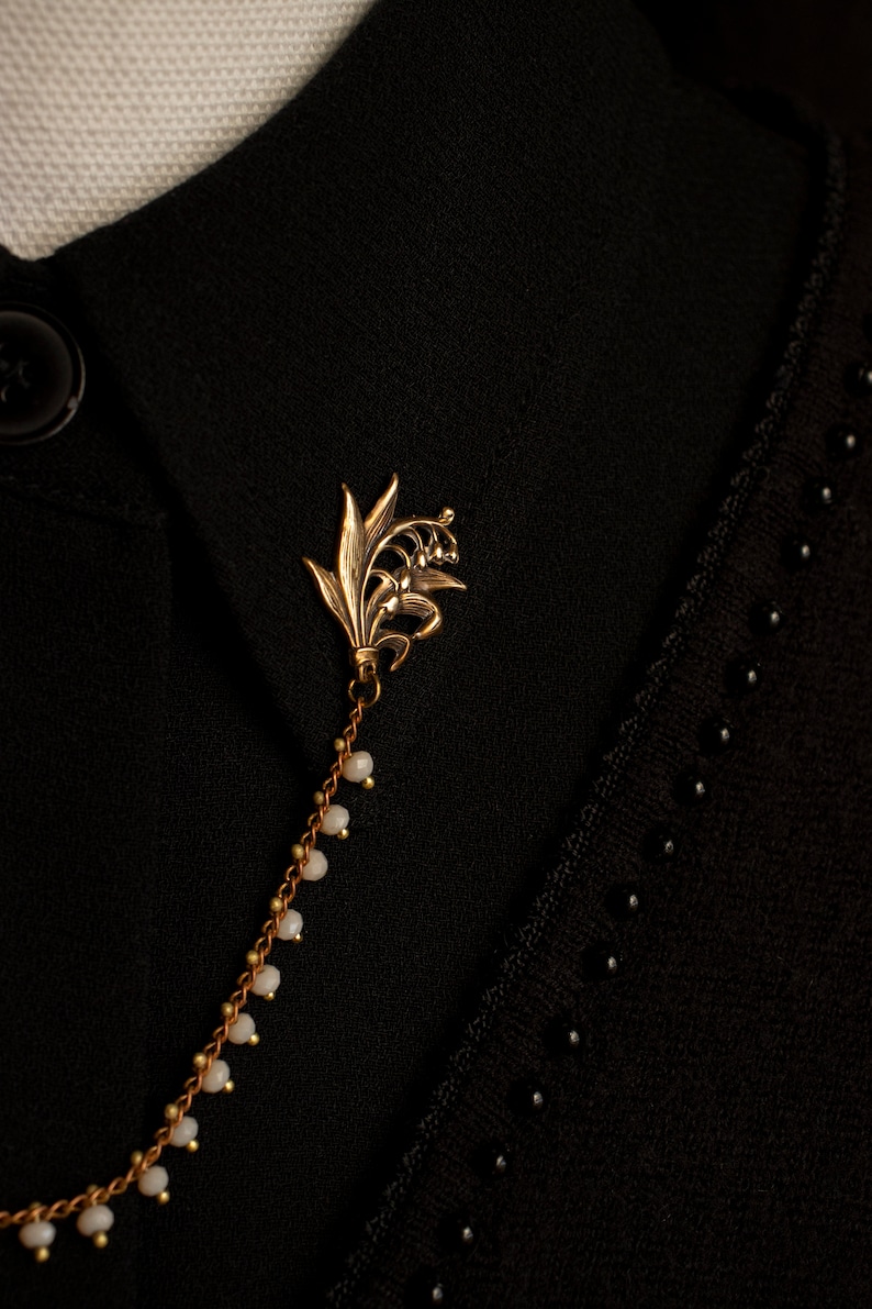 May include: A gold lily of the valley brooch with a chain of small white beads. The brooch is pinned to a black fabric background.
