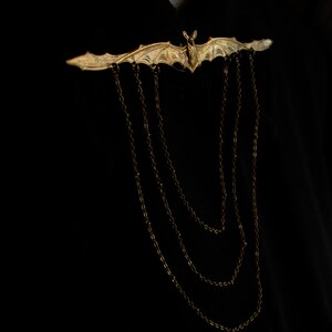 Pu&ograve; includere: Un fermaglio a forma di pipistrello dorato con un dettaglio a catena. Il fermaglio &egrave; indossato su una camicia nera.