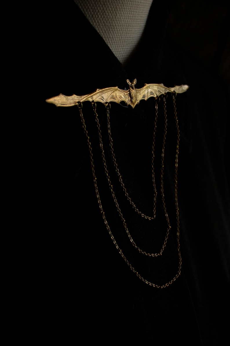 Pu&ograve; includere: Un fermaglio a forma di pipistrello dorato con un dettaglio a catena. Il fermaglio &egrave; indossato su una camicia nera.