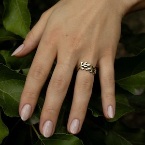 May include: A gold-colored ring with a leaf design is worn on a finger. The ring is the focal point, set against a backdrop of green leaves. The hand has polished nails.
