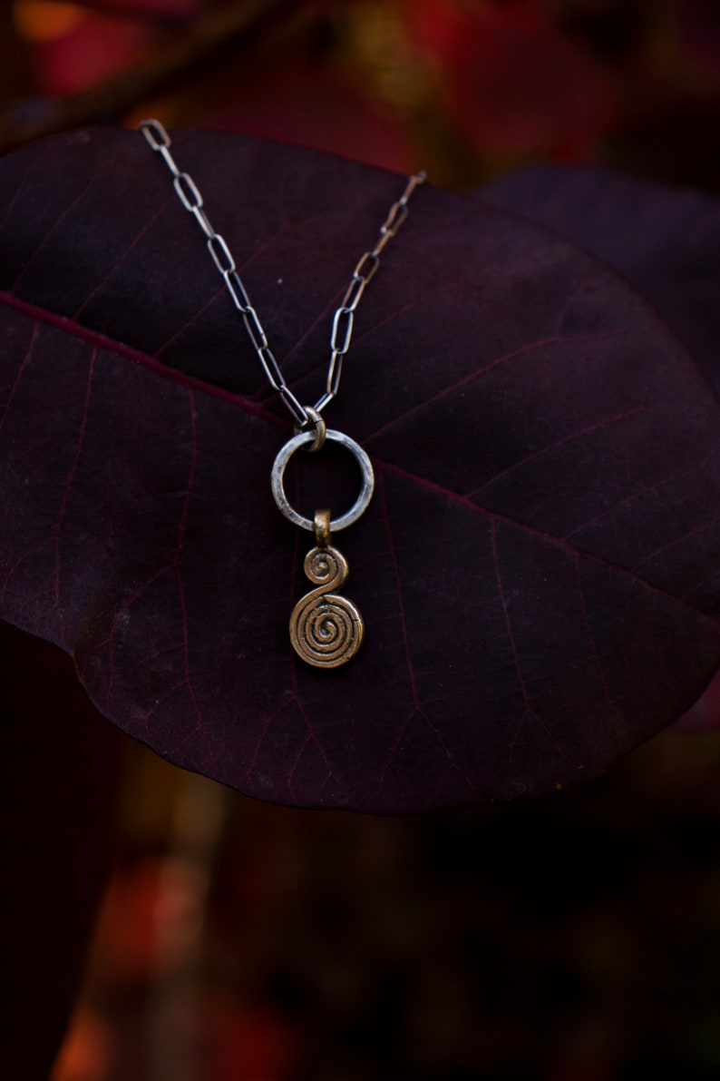 Puede incluir: Un collar de cadena de plata con un colgante circular y un encanto en espiral. El collar est&aacute; descansando sobre una hoja de color p&uacute;rpura oscuro.